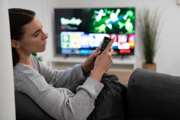 a woman sitting on a couch looking at her cell phone streaming platforms, digital overload, tv watching