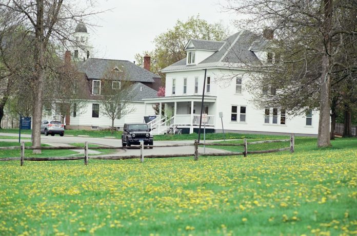 White houses and cars on a street with green field. quiet street, tree lined, small houses