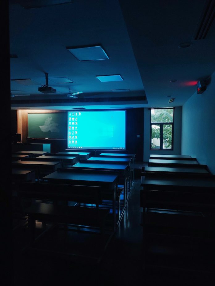 a dark room with tables and chairs and a projector screen teacher laptop dashboard charts classroom