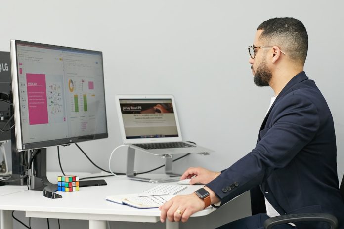 a man sitting at a desk with a laptop and a computer business automation dashboard, data flow visualization, enterprise software interface