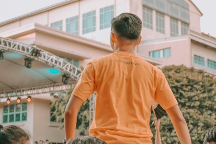 man in orange crew neck t-shirt sitting on gray rock during daytime school community event, parents teachers gathering, modern school campus