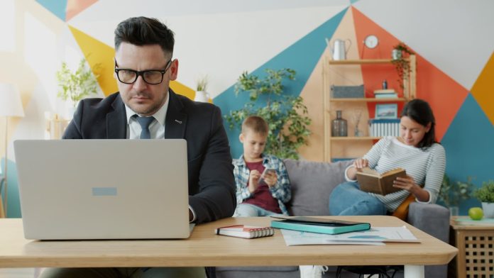 Man working on laptop while family relaxes nearby teacher parent video call, laptop meeting, home office setting