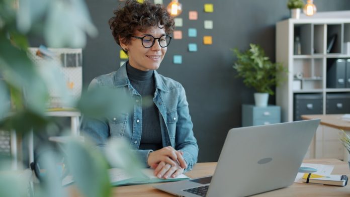 A smiling woman with curly hair works at a laptop. remote developers video call, laptop screen meeting, distributed team collaboration, home office workspace