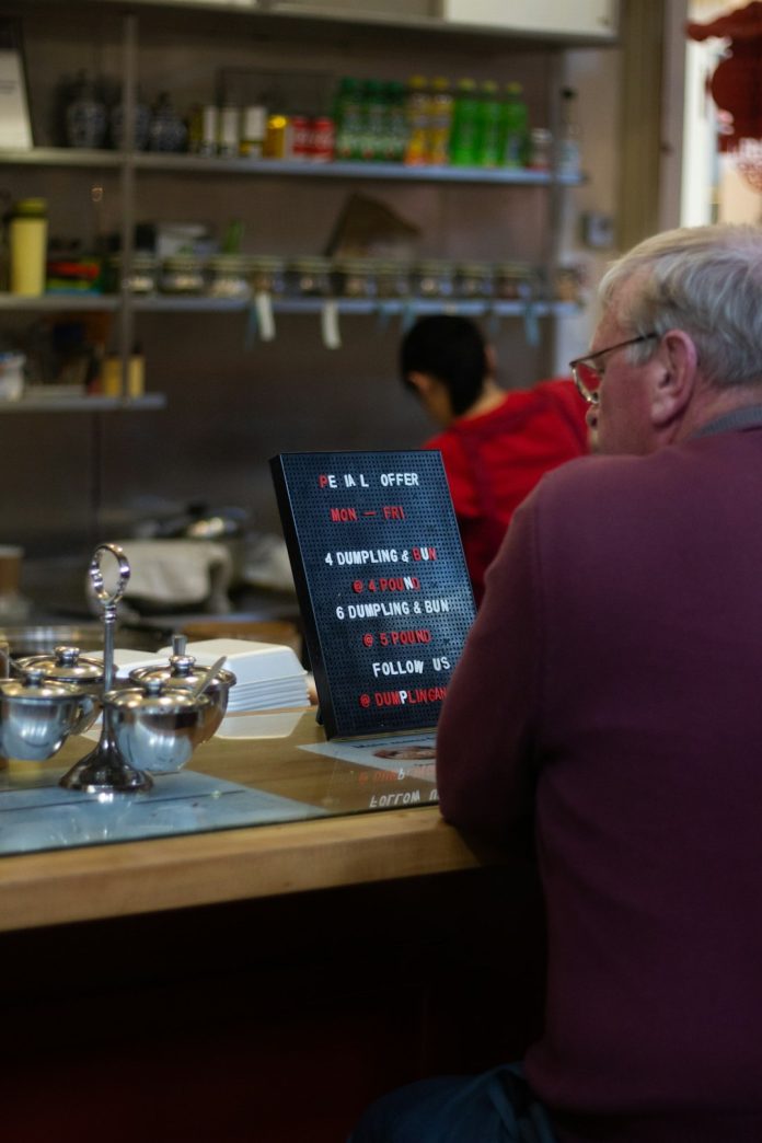 man in red sweater sitting beside brown wooden table restaurant kitchen display screen, chefs cooking, digital order system