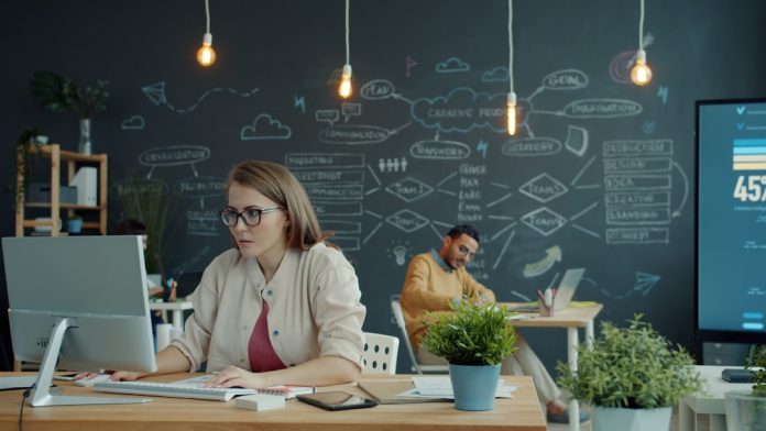Woman working on computer in modern office with chalkboard. IT service management dashboard, enterprise workflow system, operations monitoring screen, modern office workspace