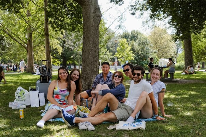 a group of people sitting on top of a lush green park friends outdoors, phones away, sunny park