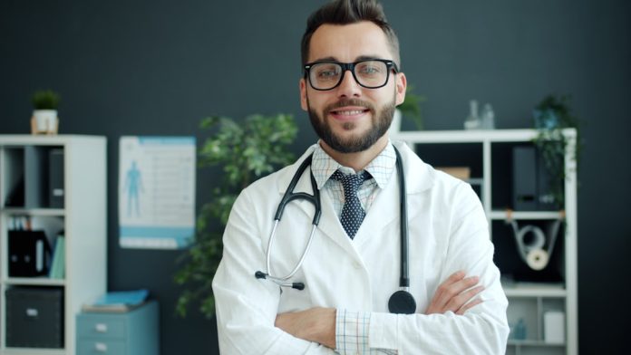 A smiling doctor with glasses and stethoscope stands arms crossed. dentist writing blog, dental education, patient trust